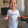 boy, cactus, carpet, casual_clothing, child, decor, floor_tiles, furniture, happy, home, indoor, living_room, monster_truck, person, portrait, shelf, shorts, smiling, toy, tshirt