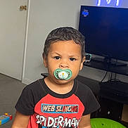 Kaiden is registered to the contest to win money with this photo: toddler, child, pacifier, spiderman_shirt, toy, indoor, carpet, television, neon_light, door, table, green_toy, yellow_truck, orange_toy, black_hair, serious_expression, person, room, furniture, playtime