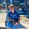 person, dog, blue_shirt, shorts, outdoor, sunny, plaza, water_feature, shade_structure, trash_bins, smiling, sitting, leash, bottle, concrete, trees, people, background, daytime, casual
