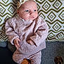 baby, infant, headband, flower, pink_sweater, patterned_pants, slippers, blanket, geometric_pattern, couch, texture, indoor, cute, child, young, portrait, cozy, warm_colors, lying_down, curious