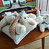 animal_toys, blue_eyes, cartoon, coffee_table, cushion, fluffy, furry, home_decor, indoors, living_room, pink_nose, plush_toy, purple_collar, stuffed_animal, television, toy_cat, toy_dog, unicorn_toy, white, wooden_furniture