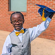 Denzel is registered to the contest to win money with this photo: achievement, boy, brick_wall, celebration, child, glasses, graduate, graduation_cap, happy, outdoor, person, portrait, smiling, standing, success, sunlight, vest, white_shirt, yellow_bow_tie, young