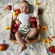 Elio participe au concours pour gagner de l'argent avec cette photo : baby, basket, apple, flower, yellow_flower, white_flower, blanket, smiling, happy, infant, clothing, shorts, white_shirt, skin, foot, hand, head, cheerful, lying_down, indoors