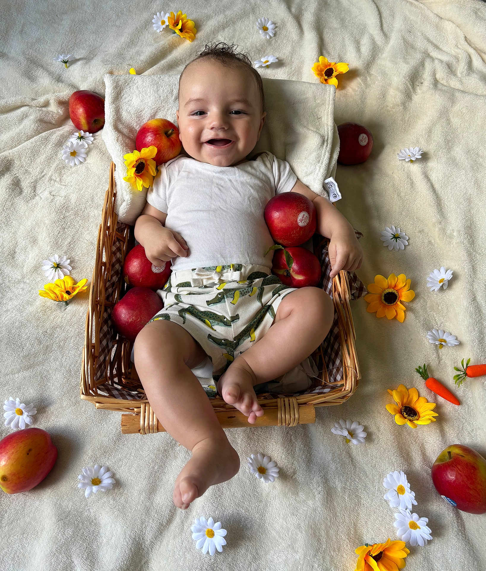 Elio participe au concours pour gagner de l'argent avec cette photo : baby, basket, apple, flower, yellow_flower, white_flower, blanket, smiling, happy, infant, clothing, shorts, white_shirt, skin, foot, hand, head, cheerful, lying_down, indoors