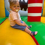 Celeyne a rejoint le concours — aidez-le/la à gagner de superbes lots ! child, toddler, pacifier, bouncy_castle, inflatable, playground, sitting, smiling, pigtails, striped_socks, pink_socks, shorts, tshirt, colorful, red_striped_pillar, yellow_floor, green_floor, blue_background, cartoon_shark, portrait