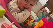 Liliana joined the competition — help win amazing prizes! baby, child, infant, smiling, sitting, activity_center, baby_toys, colorful_toys, highchair, playtime, indoor, portrait, face, hands, brown_hair, long_sleeves, pink, orange, plastic, home