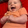 Liliana is registered to the contest to win money with this photo: baby, blurred, casual, child, cute, face, happy, head, indoor, infant, joyful, lying_down, orange_clothing, person, plaid_blanket, red_background, small_hands, smiling, toddler, young_child