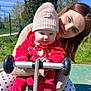 baby, woman, child, outdoor, playground, hat, red_clothing, smile, grass, fence, sunny, daylight, person, play_equipment, holding, nature, happy, park, parent, support