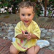 Taline participe au concours pour gagner de l'argent avec cette photo : toddler, child, curly_hair, yellow_shirt, smiling, sitting, outdoor, stone_floor, brick_floor, greenery, cute, person, happy, casual_clothing, small_object, shoes, playful, nature, portrait, daylight