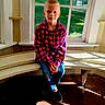 child, boy, portrait, sitting, window, indoors, sunlight, wooden_floor, bench, plaid_shirt, red_shirt, blue_jeans, shoes, blond_hair, smiling, face, home, window_seat, natural_light, photography
