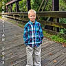 Sebastian is registered to the contest to win money with this photo: child, boy, portrait, outdoor, bridge, footbridge, wooden_bridge, forest, woodland, nature, standing, smiling, blond_hair, plaid_shirt, button_up_shirt, trousers, hands_in_pockets, rustic, photography, text_overlay