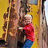 child, boy, smiling, outdoor, portrait, industrial, machinery, ladder, rust, peeling_paint, yellow, red_shirt, blue_jeans, blond_hair, climbing, adventure, railway, vehicle, weathered, photography