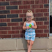 Hermione joined the competition — help win amazing prizes! child, girl, toddler, smiling, glasses, blonde_hair, ponytail, portrait, standing, outdoor, brick_wall, jumpsuit, striped_clothing, blue_outfit, crocs, decorated_shoes, snack_packet, fashion, daylight, full_body