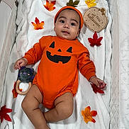 Andrés is registered to the contest to win money with this photo: baby, infant, orange_clothing, halloween, pumpkin_face, plush_toy, white_blanket, autumn_leaves, wooden_sign, cute, lying_down, indoors, headwear, smiling, child, celebration, seasonal, festive, soft_texture, small_hand