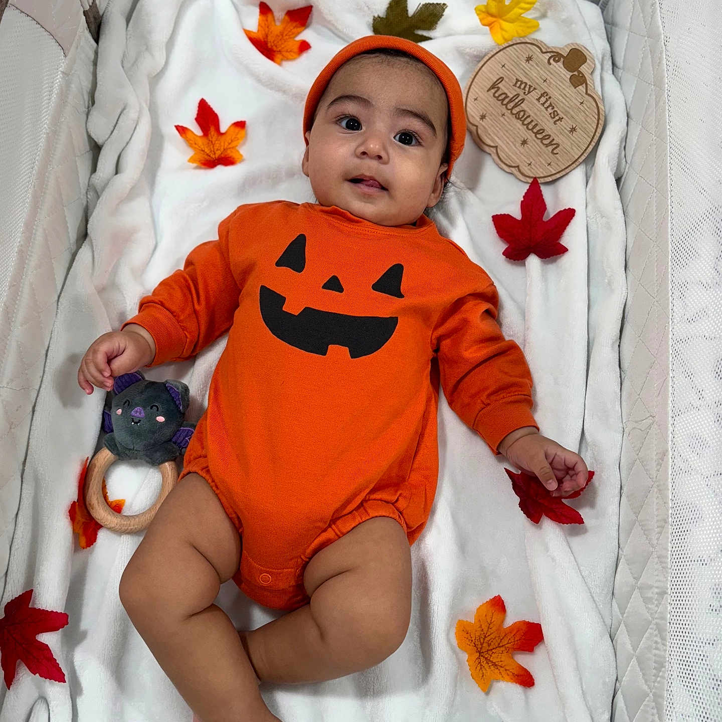 Andrés is registered to the contest to win money with this photo: autumn_leaves, baby, celebration, child, cute, festive, halloween, headwear, indoors, infant, lying_down, orange_clothing, plush_toy, pumpkin_face, seasonal, small_hand, smiling, soft_texture, white_blanket, wooden_sign