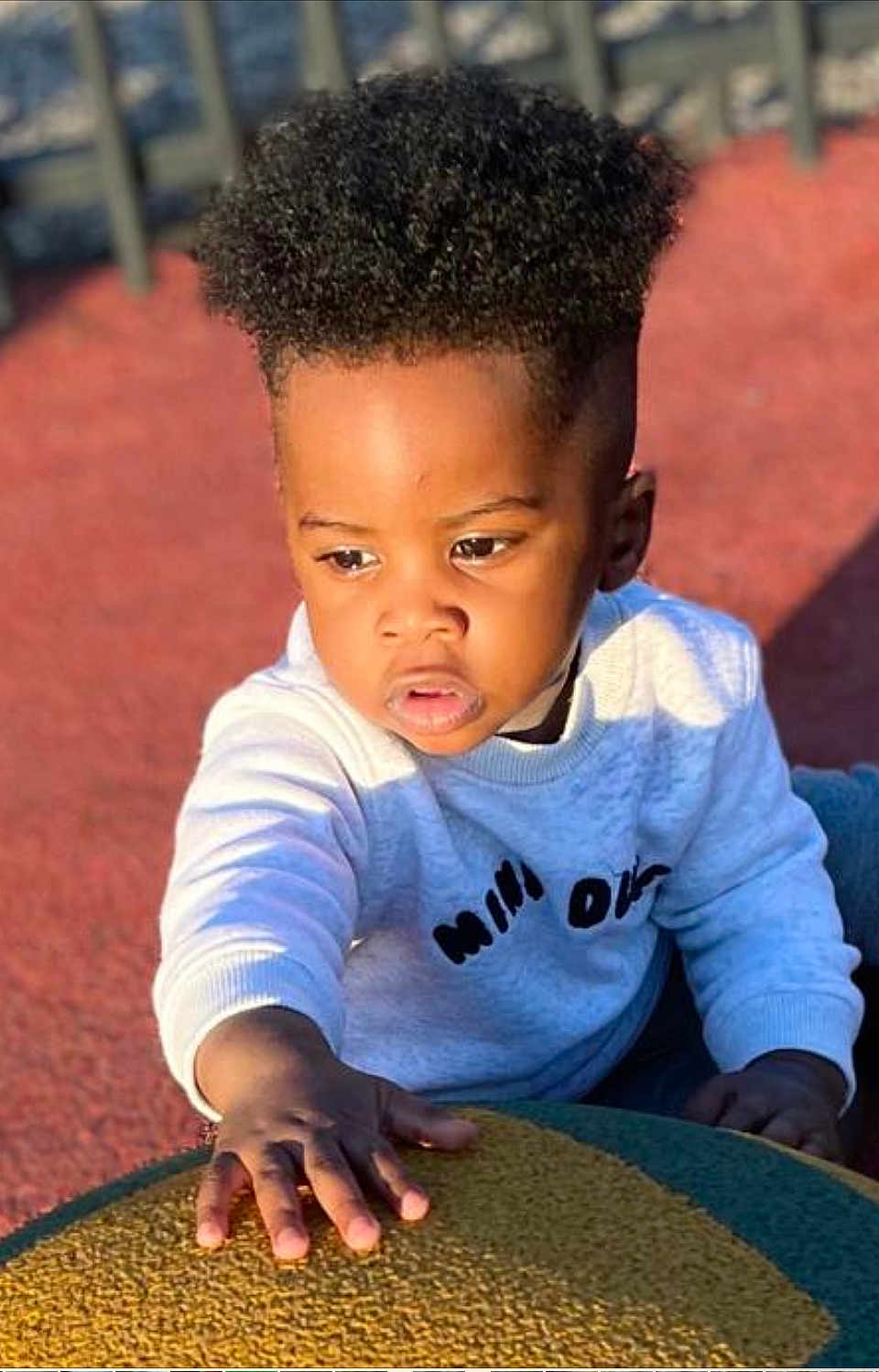 Grâce Miguel participe au concours pour gagner de l'argent avec cette photo : child, toddler, curly_hair, sweater, outdoor, playground, sunlight, hand, face, expression, texture, colorful, young, person, casual_clothing, daylight, skin, portrait, activity, curiosity