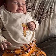 Teddy is registered to the contest to win money with this photo: baby, child, infant, smiling, cute, bib, winnie_the_pooh, tigger, orange_pants, couch, cushions, indoor, clothing, portrait, face, hand, relaxed, home, toddler, person