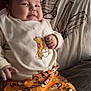 baby, child, infant, smiling, cute, bib, winnie_the_pooh, tigger, orange_pants, couch, cushions, indoor, clothing, portrait, face, hand, relaxed, home, toddler, person