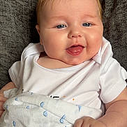 Teddy is registered to the contest to win money with this photo: baby, child, smiling, blanket, white_clothing, blue_pattern, face, tongue_out, hand, cushion, indoor, comfort, cute, happy, infant, portrait, skin, head, person, seated