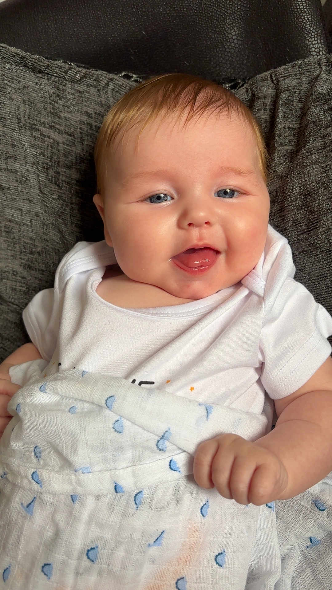 Teddy is registered to the contest to win money with this photo: baby, child, smiling, blanket, white_clothing, blue_pattern, face, tongue_out, hand, cushion, indoor, comfort, cute, happy, infant, portrait, skin, head, person, seated