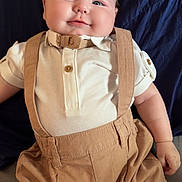 Teddy is registered to the contest to win money with this photo: baby, beige, blue, bow_tie, chubby, clothing, cute, face, happy, indoors, infant, person, pillow, portrait, seated, shorts, skin, smile, suspenders, young_child