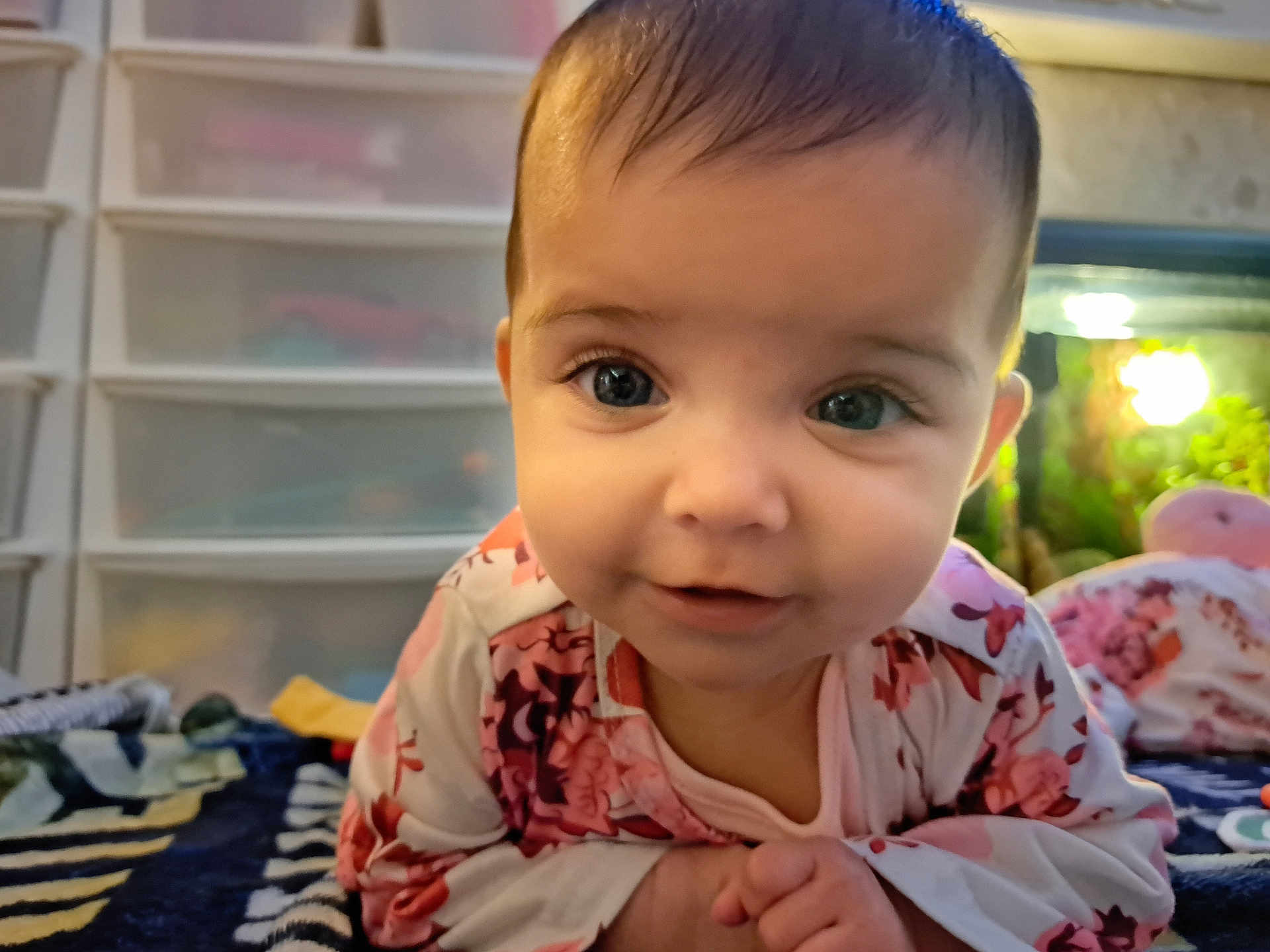 Neveah is registered to the contest to win money with this photo: baby, child, infant, face, eyes, floral_clothing, lying_down, hands, curious, indoors, soft_light, background_blur, fish_tank, toy, blanket, cute, portrait, close_up, home, sleepwear