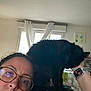 woman, glasses, black_cat, cat, indoor, window, curtain, handprint_art, watch, face, person, shoulder, home, casual, portrait, pet, furniture, wall, natural_light, relaxed