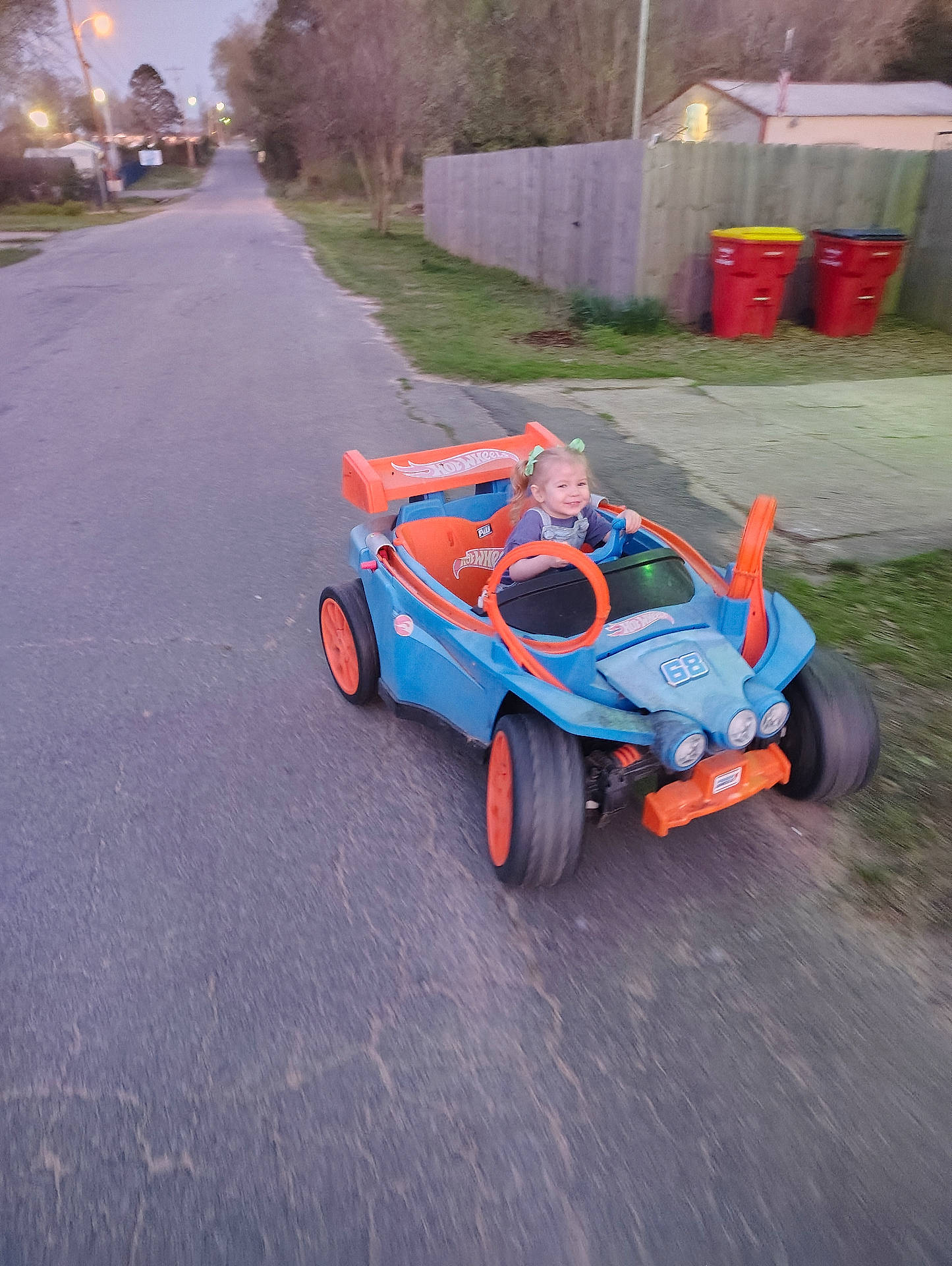 Jalynn joined the competition — help win amazing prizes! blue_car, child, driveway, evening, fence, grass, happy, headlights, motion_blur, orange_wheels, pavement, plastic_toy, road, smile, steering_wheel, street, suburban, toddler, toy_car, trash_bins