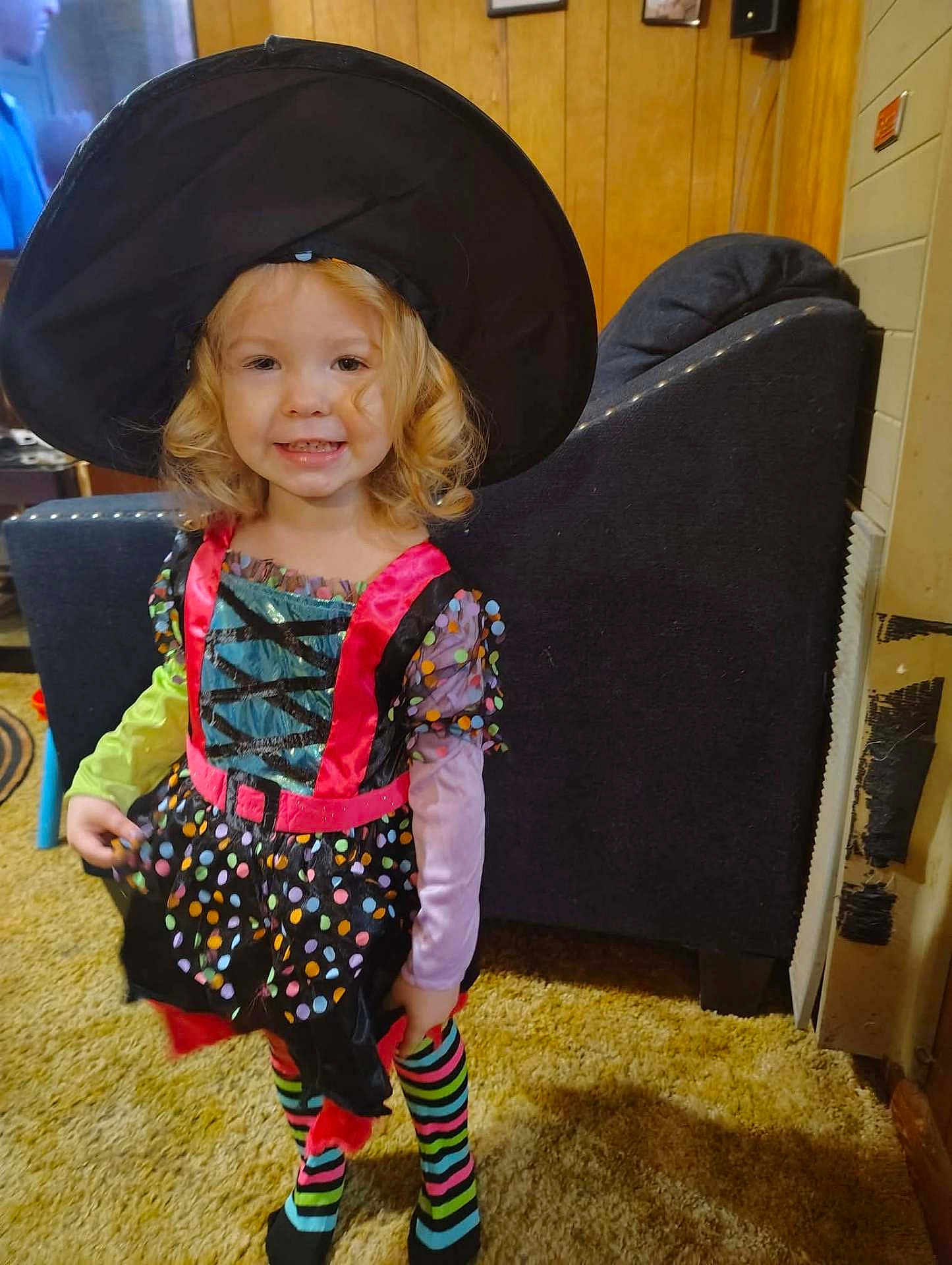 Jalynn is registered to the contest to win money with this photo: child, toddler, girl, smile, costume, witch_hat, striped_socks, indoor, living_room, carpet, chair, wood_paneling, curly_hair, portrait, smiling, happy, playful, furniture, television, hat