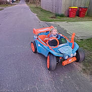Jalynn is registered to the contest to win money with this photo: blue_car, child, dusk, grass, happy, headlights, motion_blur, neighborhood, orange_wheels, outdoor_play, pavement, ride_on_toy, smile, steering_wheel, street, toddler, toy_car, trash_bins, wheels, wooden_fence