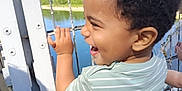 Tyler participe au concours pour gagner de l'argent avec cette photo : child, toddler, smiling, laughing, curly_hair, striped_shirt, outdoor, daylight, metal_fence, river, water, trees, nature, greenery, happy, holding, casual_clothing, profile, young_child, sunny_day