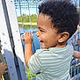 child, toddler, smiling, laughing, curly_hair, striped_shirt, outdoor, daylight, metal_fence, river, water, trees, nature, greenery, happy, holding, casual_clothing, profile, young_child, sunny_day