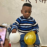 Michel a rejoint le concours — aidez-le/la à gagner de superbes lots ! toddler, child, balloon, blue_striped_sweater, white_balloon, golden_balloon, sitting, chair, indoor, person, smartphone, photography, celebration, party, casual_clothing, young_child, curly_hair, neutral_expression, hand, footwear