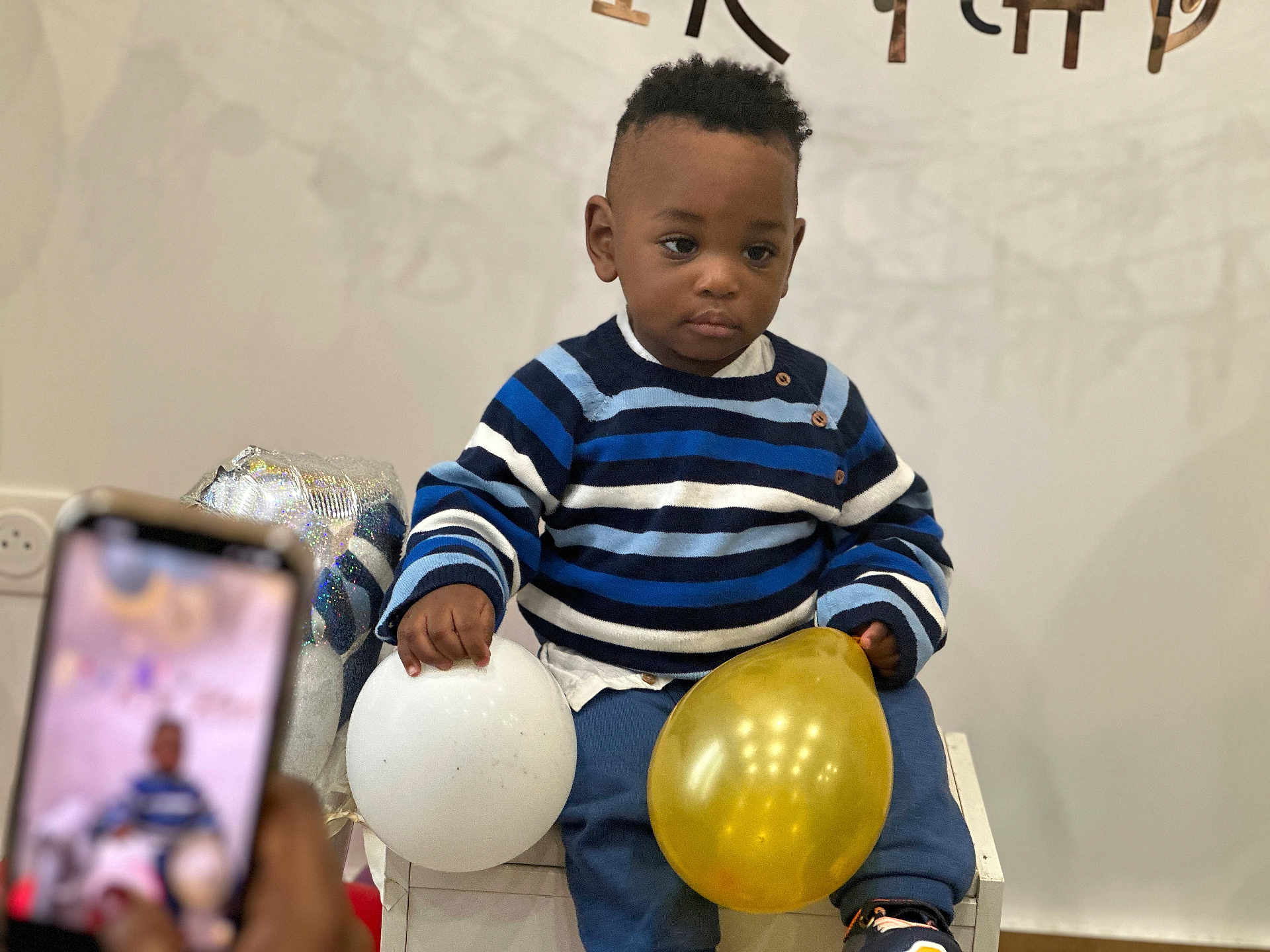 Michel a rejoint le concours — aidez-le/la à gagner de superbes lots ! toddler, child, balloon, blue_striped_sweater, white_balloon, golden_balloon, sitting, chair, indoor, person, smartphone, photography, celebration, party, casual_clothing, young_child, curly_hair, neutral_expression, hand, footwear