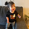 child, toddler, baby, smiling, happy, standing, portrait, indoors, living_room, couch, sofa, striped_leggings, black_shirt, ghost_design, halloween, pigtails, warm_lighting, houseplant, home_decor, photography