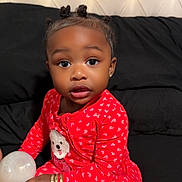 Skylar is registered to the contest to win money with this photo: child, toddler, baby, portrait, sitting, bed, black_bedding, tufted_headboard, red_outfit, heart_pattern, pajamas, hair_buns, earrings, bracelets, toy, ball, indoors, bedroom, looking_at_camera, person