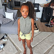 Awa a rejoint le concours — aidez-le/la à gagner de superbes lots ! child, girl, living_room, barefoot, carpet, curly_hair, high_ponytail, green_shirt, yellow_shorts, modern_kitchen, sofa, pillows, slippers, wooden_floor, bracelets, casual_clothing, indoor, person, standing, home