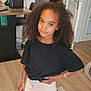 child, girl, barefoot, curly_hair, black_shirt, shorts, bracelet, living_room, rug, wooden_floor, door, coat_hanger, bag, table, chair, earrings, smile, standing, indoor, casual