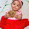Rya is registered to the contest to win money with this photo: baby, child, tutu, red_skirt, pink_headband, holding_sign, wooden_sign, ten_months, cute, smiling, infant, portrait, lying_down, colorful_blanket, young_child, clothing, face, hands, indoor, person