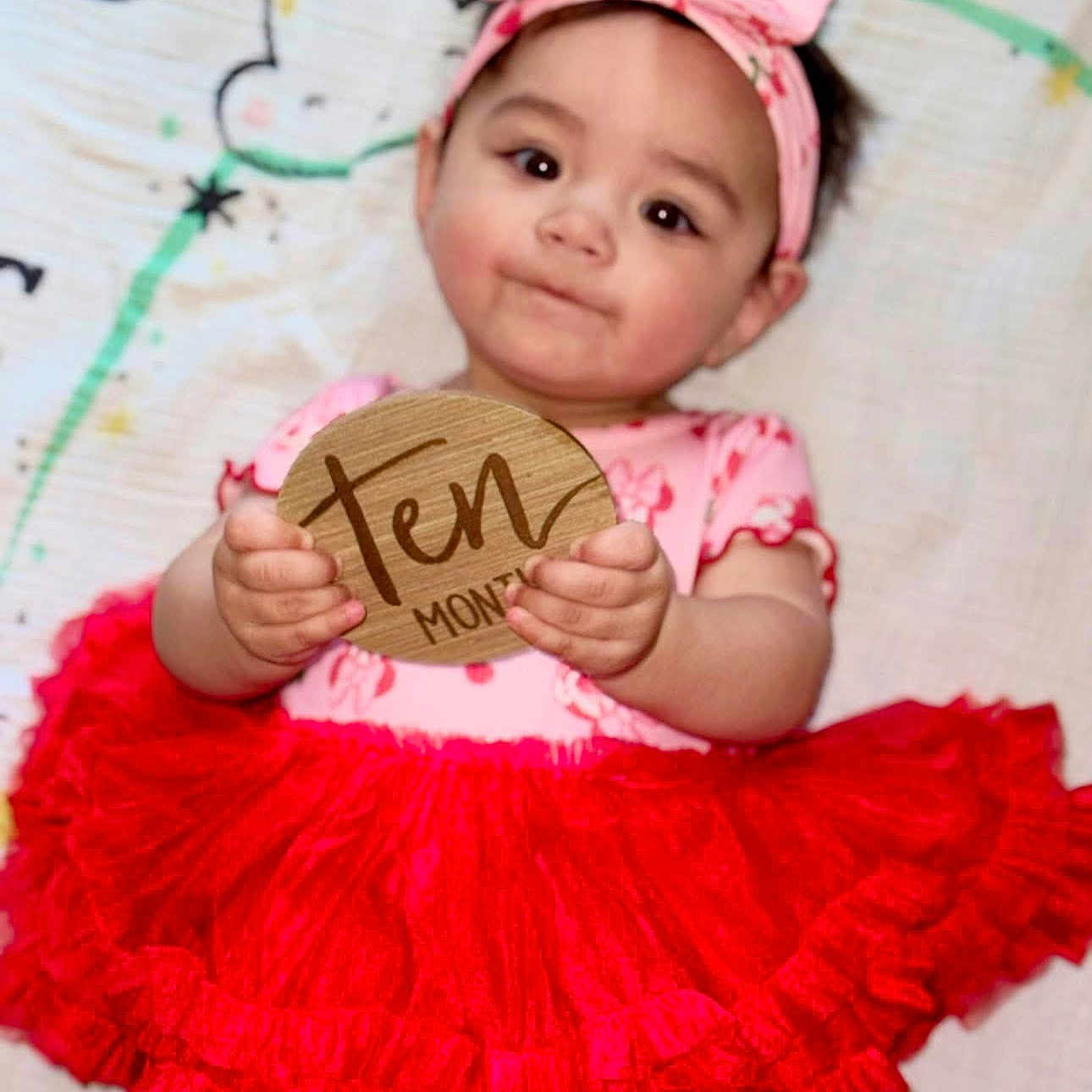 Rya is registered to the contest to win money with this photo: baby, child, tutu, red_skirt, pink_headband, holding_sign, wooden_sign, ten_months, cute, smiling, infant, portrait, lying_down, colorful_blanket, young_child, clothing, face, hands, indoor, person