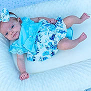 Ember is registered to the contest to win money with this photo: baby, infant, child, blue_dress, headband, bow, floral_pattern, cushion, white_bedding, lying_down, cute, portrait, smile, skin, person, newborn, soft_texture, indoors, relaxed, baby_girl