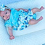 baby, infant, child, blue_dress, headband, bow, floral_pattern, cushion, white_bedding, lying_down, cute, portrait, smile, skin, person, newborn, soft_texture, indoors, relaxed, baby_girl