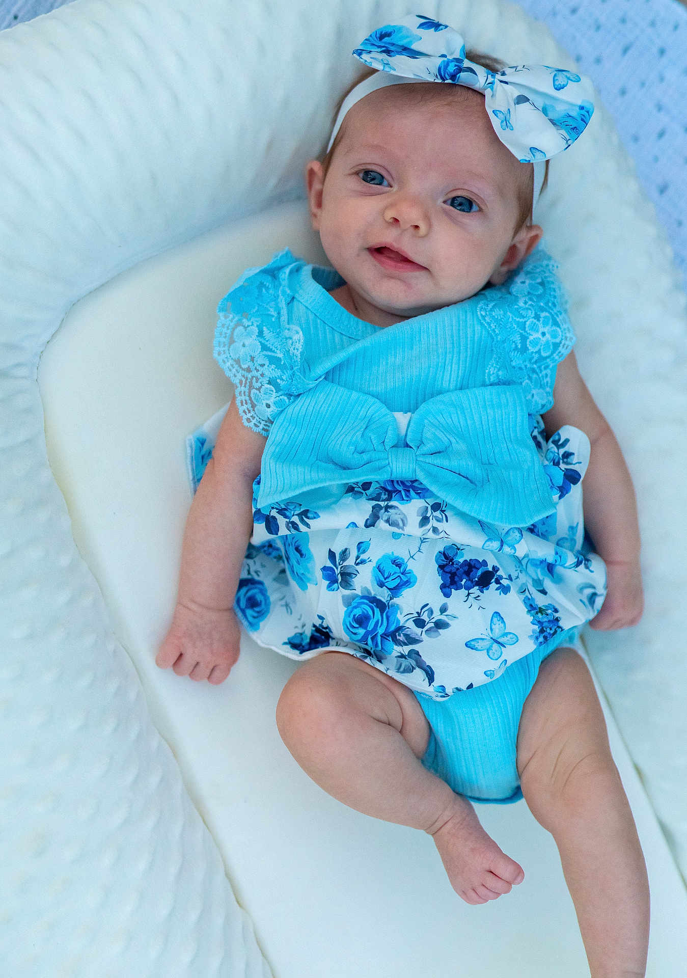 Ember joined the competition — help win amazing prizes! baby, infant, blue_clothing, bow, headband, floral_pattern, smile, cute, lying_down, soft_cushion, white_background, portrait, child, skin, tiny_hands, tiny_feet, happy, eyes, indoors, comfortable