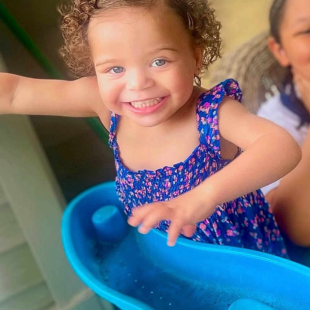 Jaelyn is registered to the contest to win money with this photo: adult, blue_dress, blue_eyes, blurry_background, bright, casual, child, curly_hair, cute, family, floral_pattern, happy, indoor, joy, person, play_structure, portrait, reaching, smiling, toddler