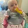 baby, banana, bib, child, countertop, cute, drool, earring, eating, floral_print, highchair, indoor, infant, kitchen, nike_logo, onesie, portrait, sofa, teething, window