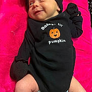 Aurora is registered to the contest to win money with this photo: baby, infant, black_clothing, mittens, socks, headwrap, smile, pink_blanket, cute, lying_down, cheeky_expression, pumpkin_graphic, cozy, newborn, infant_fashion, portrait, child, human, soft_texture, indoors