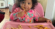 Kaylee joined the competition — help win amazing prizes! toddler, child, high_chair, food, french_fries, snack, curly_hair, pink, onesie, indoor, home, table, seat, eating, person, cute, baby, face, expression, furniture
