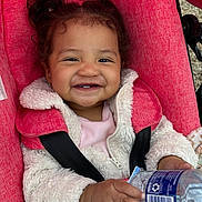 Kaylee is registered to the contest to win money with this photo: toddler, child, smiling, face, jacket, stroller, red, water_bottle, hand, outdoor, happy, person, baby, cute, clothing, seatbelt, portrait, curly_hair, cozy, playful