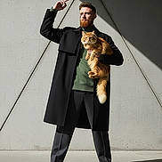 Ms.kitty is registered to the contest to win money with this photo: man, cat, orange_cat, red_beard, black_coat, green_sweater, gray_pants, black_boots, concrete_wall, shadow, sunlight, standing, posing, outdoor, fashion, pet, fluffy_cat, portrait, confident, urban