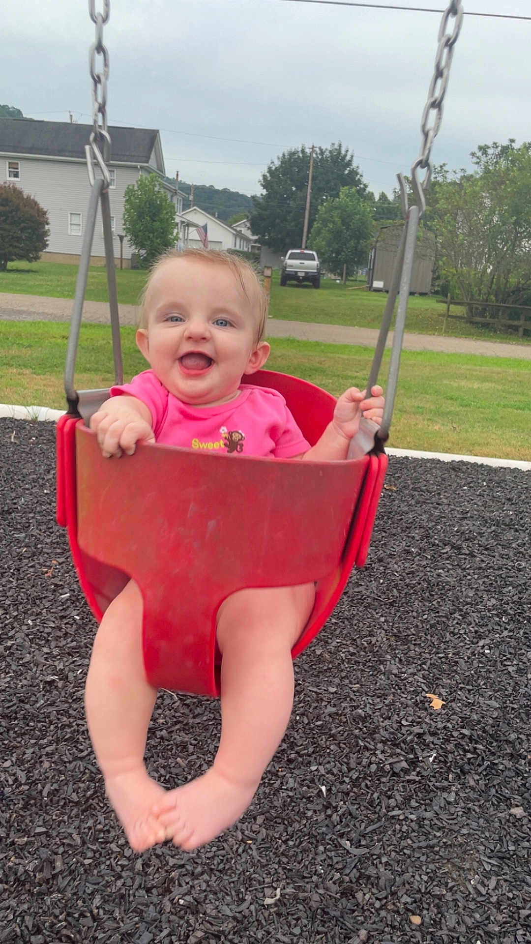 Rosalie joined the competition — help win amazing prizes! baby, child, swing, playground, red_swing, pink_shirt, smiling, happy, outdoor, grass, trees, houses, residential_area, feet, legs, summer, daytime, fun, park, person